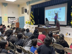 Photograph of a school auditorium presentation showing a speaker pointing at a projected map or diagram on a screen, with two other individuals seated nearby. Audience of students seated in rows faces the stage, with decorations including yellow and black balloons and a "MUSIC" sign visible on the left side.