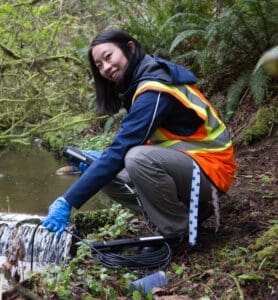 A photograph of a person wearing a high-visibility safety vest and gloves, crouching near a small stream in a forested area while handling water monitoring equipment. The scene suggests fieldwork or environmental data collection related to water or ecological monitoring.