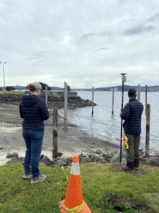 Two people standing near a shoreline, each holding surveying equipment, conducting a land or water measurement. The scene includes a grassy area with an orange traffic cone in the foreground and a cloudy sky over a body of water with rocks and wooden posts along the shore.